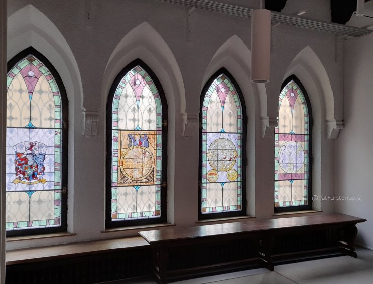 Four tall, pointed Gothic-style window arches, stained glass windows displaying historical maps, star signs, and coats of arms.